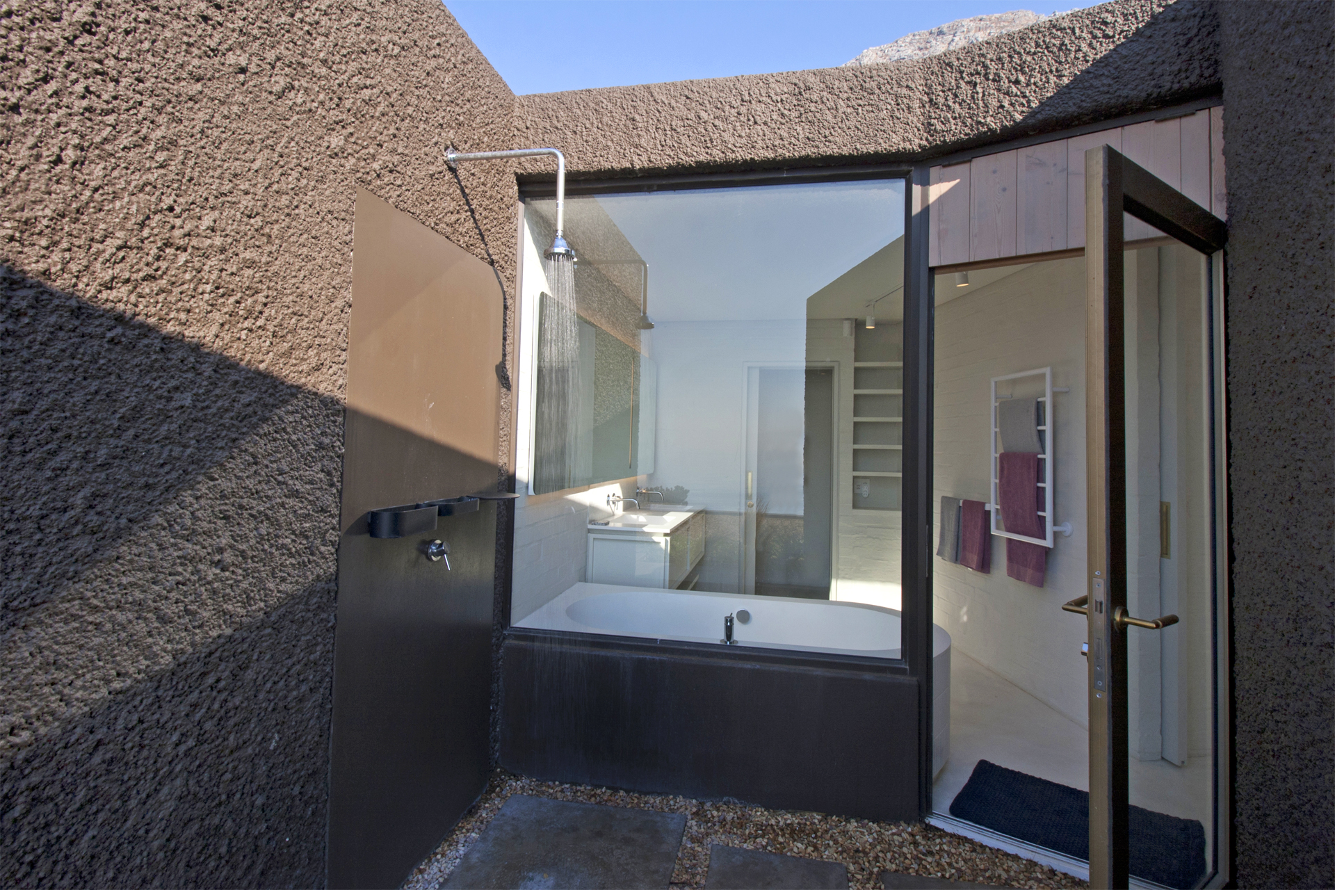Private outdoor shower area with a view into a modern bathroom featuring a bathtub and large windows.